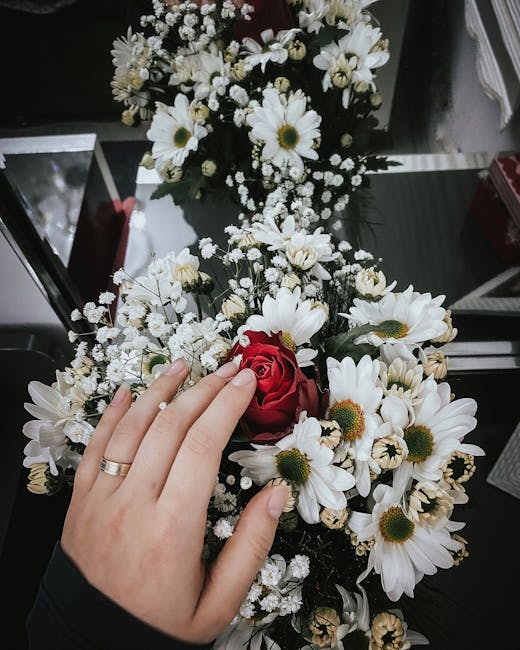 A florist's hand with a silver band on the ring finger is gently holding a small bouquet of fresh flowers, consisting of a single red rose and several white daisies with yellow centers, surrounded by
