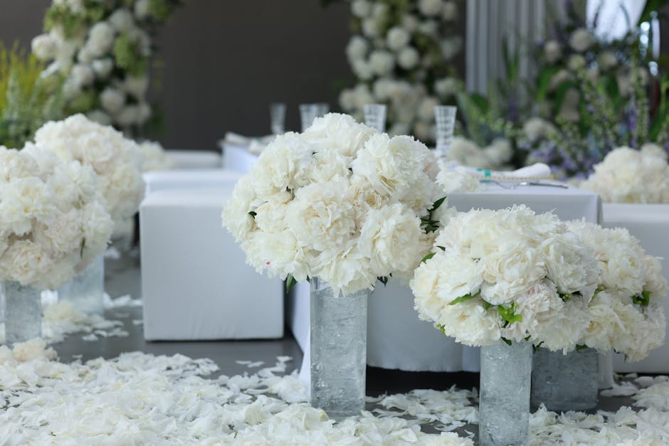 A floral arrangement featuring large, lush bouquets of white peonies and hydrangeas in tall, rectangular glass vases, placed on a white surface sprinkled with white flower petals. In the background, t