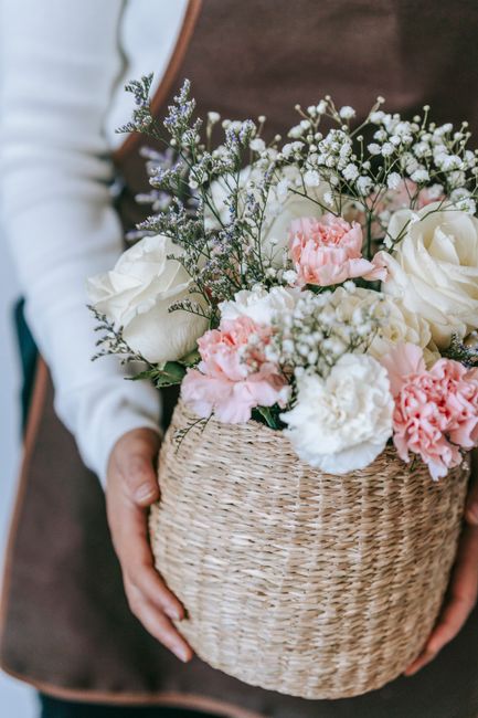 A person holding a woven wicker basket filled with a fresh floral arrangement consisting of white roses, soft pink carnations, and delicate sprigs of baby's breath and lavender. The flowers are vibran