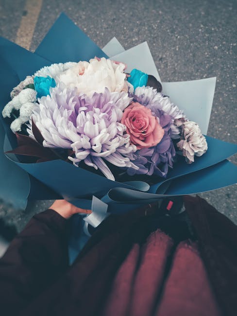 A hand holding a vibrant bouquet of fresh flowers wrapped in blue and white paper, including a pink rose, white chrysanthemums, purple asters, and bright blue accents. The arrangement is lush and care