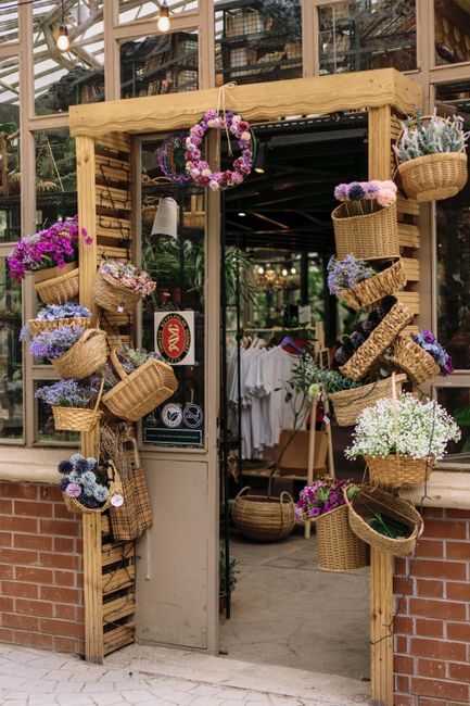 The entrance to a flower shop features a rustic wooden archway adorned with hanging baskets and decorative floral wreaths, showcasing an array of colourful flowers such as purple, white, and pink bloo