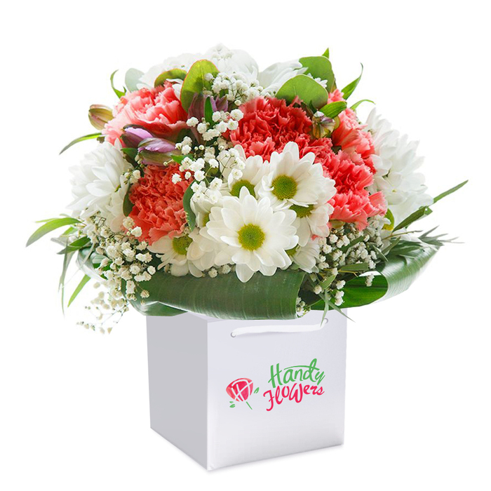 A visually arresting bouquet, upright in a clean white gift bag, captures the grace and colour of Old Malden's flower-rich landscapes. The centrepiece features coral-pink and subtle peach carnations, their distinctive ruffled petals creating depth and a sense of plush opulence, interspersed with pristine white daisies that show vivid chartreuse centers-each bloom bursting like the first day of summer at Malden Manor Park. Threads of white baby's breath meander among the primary blooms, adding dimension and a feathery softness reminiscent of early morning dew on local hedgerows. Deep, lustrous green foliage encircles the base of the arrangement, broad glossy leaves forming a natural, elegant wrap that highlights nature's abundance. Punctuating the design, purple-kissed unopened buds on the bouquet's left introduce intrigue and gentle anticipation, much like the promise of blooms in Old Malden's hidden gardens. Finished with careful attention to balance, the bouquet forms a full, rounded silhouette that radiates freshness, artistry, and life. The bag's white ribbon handles gleam unobtrusively at the top, making the bouquet as easy to carry as it is beautiful to behold-ideal for same-day delivery across Old Malden for birthdays, anniversaries, or heartfelt gestures of appreciation. Every element reflects the skill and warmth of an experienced local florist, perfectly capturing the area's welcoming charm.